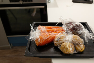 Fresh vegetables, wrapped in plastic, are arranged on a tray ready for baking. The kitchen is...