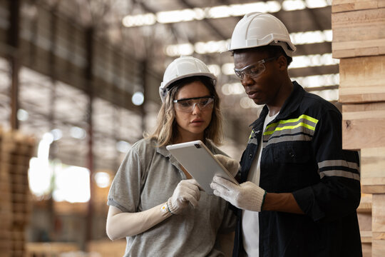 Two Multi-ethnic industrial workers wearing safety helmets and gloves use digital tablet for data analysis in warehouse, showcasing teamwork, technology wooden manufacturing processes. - Powered by Adobe