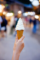A close-up of a hand holding a soft serve ice cream cone against a blurry background of a charming street in Mae Kampong, Thailand, representing a local travel experience.

