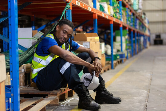 Exhausted warehouse worker in safety vest sitting on pallet with eyes closed, holding helmet, depicting fatigue, burnout, or emotional stress in logistics and industrial work environments.