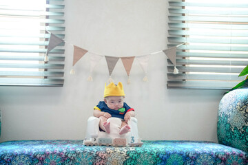 A baby wearing a crown sits on a bench with a banner hanging behind him. The banner has flags on it...