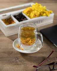 Herbal tea in clear glass mug on wooden table with floral decorations and work materials