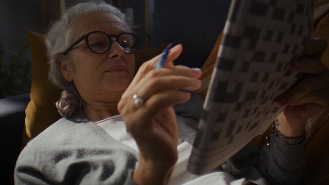 Relaxed senior woman in eyeglasses solves crossword at home