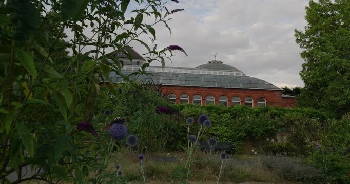 4K pan across Avery Hill Park revealing the historic Victorian Winter Garden with red brick facade, lush flowers, and Edwardian estate grounds in south east London.