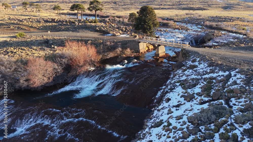 Poster drone forward descending shot toward a small bridge over a river with a tiny cascade, snow patches, 