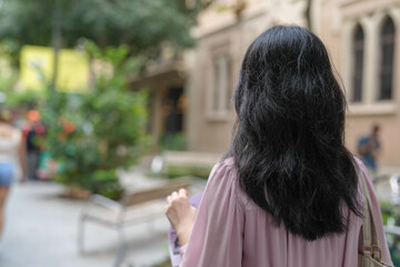 A woman with long black hair is walking down a street. She is wearing a pink shirt and carrying a purse. The scene is set in a city with a mix of people and buildings