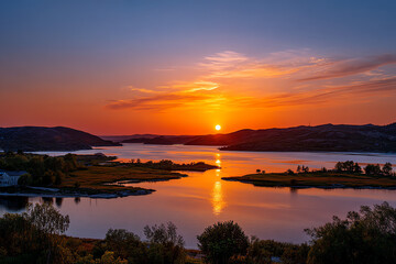 Fototapeta premium A vivid sunset over a calm lake surrounded by hills and greenery, reflecting intense orange and blue hues on the water.