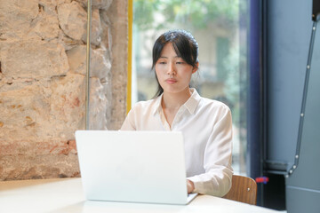 A woman is sitting at a table with a laptop in front of her. She is wearing a white shirt and has...