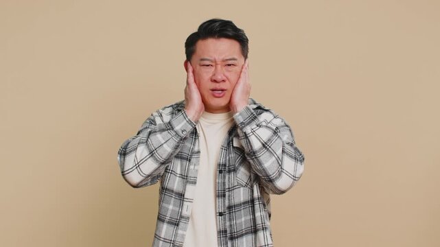 Don't want to hear, listen. Frustrated annoyed irritated Asian man covering ears gesturing no, avoiding advice ignoring unpleasant noise loud voices. Chinese mature guy isolated on beige background