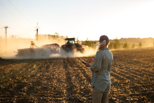 Farmer with smartphone monitoring smart farming cultivation while tractor and seeder prepare soil during agricultural work on field - Powered by Adobe