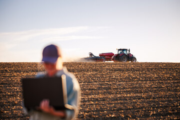 Smart farming and modern agriculture with farmer using laptop while tractor with seeder sowing crop seeds on field. Agronomist with computer analyzing farm data © encierro