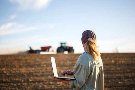 Smart farming. Woman farmer with laptop analyzing agricultural data in front of tractor and seeding machine on cultivated field - Powered by Adobe