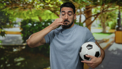 Young man with a beard holding a soccer ball making a gesture in a sunny outdoor park setting displaying a thoughtful expression.