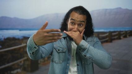 Man in denim jacket covering mouth with hand on seaside promenade street with ocean and railing...