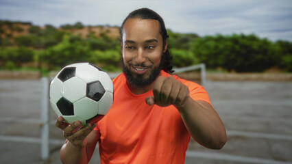 Man with beard holding a soccer ball and waving open hand while smiling on street with goalpost nearby; joy teamwork camaraderie.