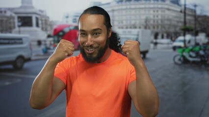 Man in bright orange shirt with fists raised pointing to chest on a busy street with buses and...