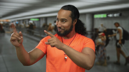 Man pointing fingers with both hands and smiling in an airport terminal wearing bright orange shirt...