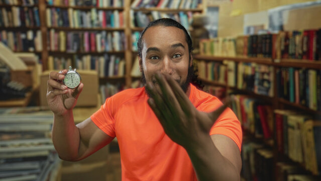 Man holds stopwatch and reaches hand forward in library building while smiling and showing stopwatch to camera; urgency time pressure.