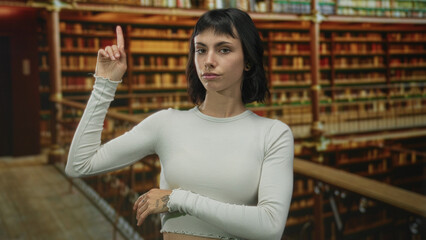 Woman with exposed hand points finger upward to wooden bookshelf railing in a library building;...