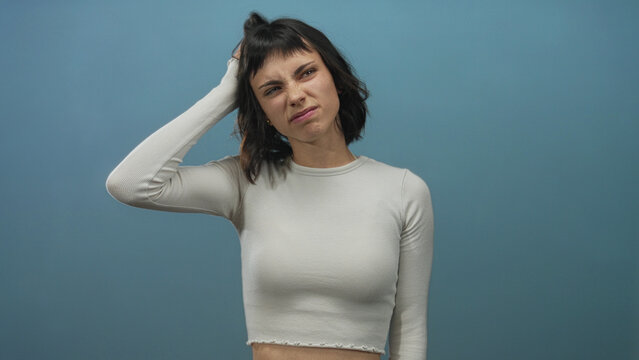 Young hispanic woman scratches head with hand in blue studio under soft light, looking puzzled; uncertainty.
