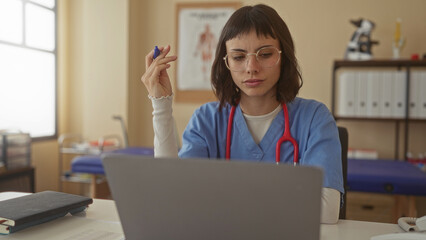 Woman holding pen at laptop in clinic with red stethoscope hanging on neck and anatomical chart...