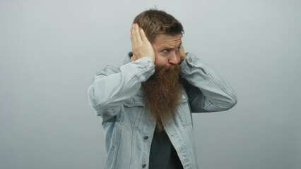 Young caucasian bearded man tightly covers ears with both hands and furrows brow in pale blue...