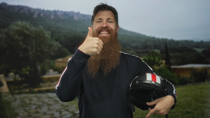 Man with beard holding helmet gives thumbs up in forest near mountain ridge and stone pathway;...