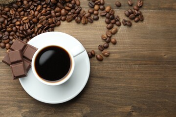 Aromatic coffee in cup, beans and pieces of chocolate on wooden table, flat lay. Space for text