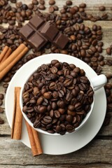 Aromatic coffee beans in cup, cinnamon and pieces of chocolate on wooden table, flat lay