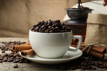 Aromatic coffee beans in cup, cinnamon and grinder on wooden table, closeup