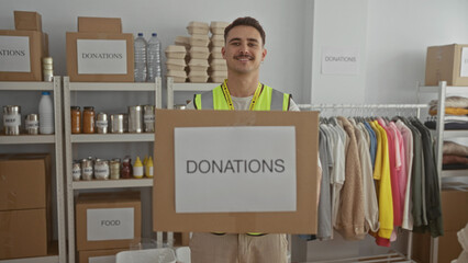 Young man in reflective vest holds donation box in charity room surrounded by shelves of clothes...