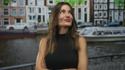 Woman with arms crossed leaning on a railing by a canal on a cobbled street among historic buildings in bright daylight; serenity.