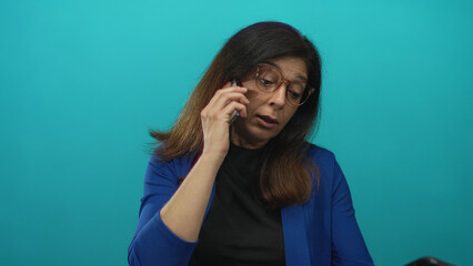 Middle aged hispanic woman with brown framed glasses wearing blue blazer and black top holds smartphone to ear in studio; concern.