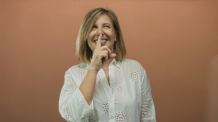 Middle aged woman pressing her index finger to lips for silence against an orange studio...