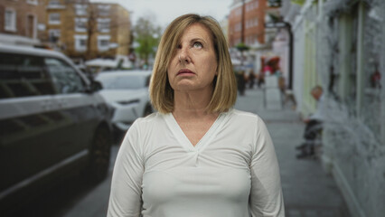 Middle aged caucasian woman purses lips in front of parked cars on busy urban street; annoyance.