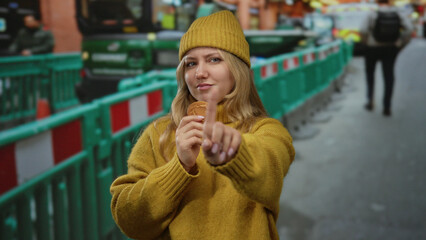 Woman in yellow sweater and beanie points finger at construction site, holding cookie, with...