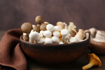 Different raw mushrooms in bowl on wooden table against brown background, closeup