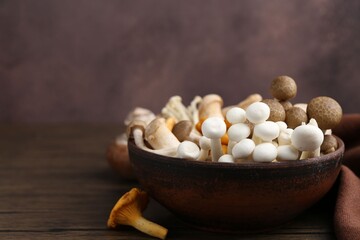 Different raw mushrooms in bowl on wooden table against brown background, closeup. Space for text