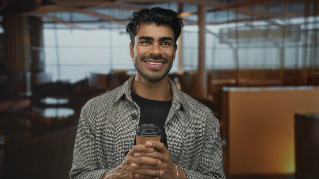 Young hispanic man smiling while holding paper coffee cup and enjoying morning beverage in modern hotel lobby; relaxation.