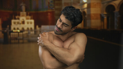 Young man wraps arms across chest in church amid blurred pews and arched columns with soft smile;...