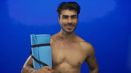 Man holding rolled yoga mat against lean bare chest in bright blue studio; confidence strength...