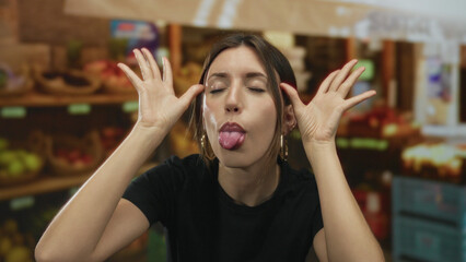 Young hispanic woman wearing black shirt makes silly face sticks out tongue and points finger at fruit in building; mischief.