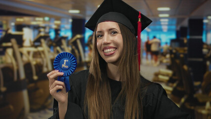 Young hispanic woman graduate wearing mortarboard and gown holds a blue first place ribbon inside...