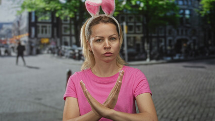 Blonde woman wearing bunny ears headband crossing arms into an x gesture on sunlit city street...