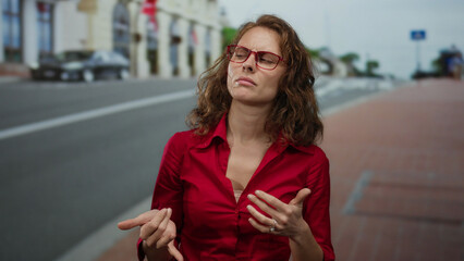 Obraz premium Woman in red shirt with glasses stands thoughtfully on a city street with buildings blurred in the background, outdoors during daytime.