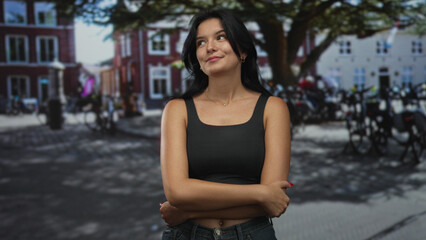 Young hispanic woman with arms crossed on sunny street wearing a black tank top and blue denim jeans; quiet confidence.