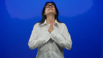 Young hispanic woman wearing glasses and striped shirt clasping hands praying in brightly lit studio; hope faith devotion.