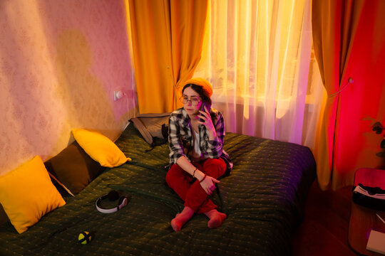 Beautiful young woman makes a voice call while sitting on bed in sunset room. Concept of leisure communication and indoor comfort. Wide shot, top view - Powered by Adobe