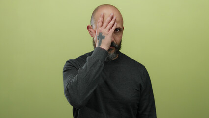 Bald man with beard and tattoo covers face on isolated yellow background conveying emotion