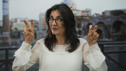 Woman wearing glasses makes air quotes gesture at roman ruins overlooking ancient colonnade;...